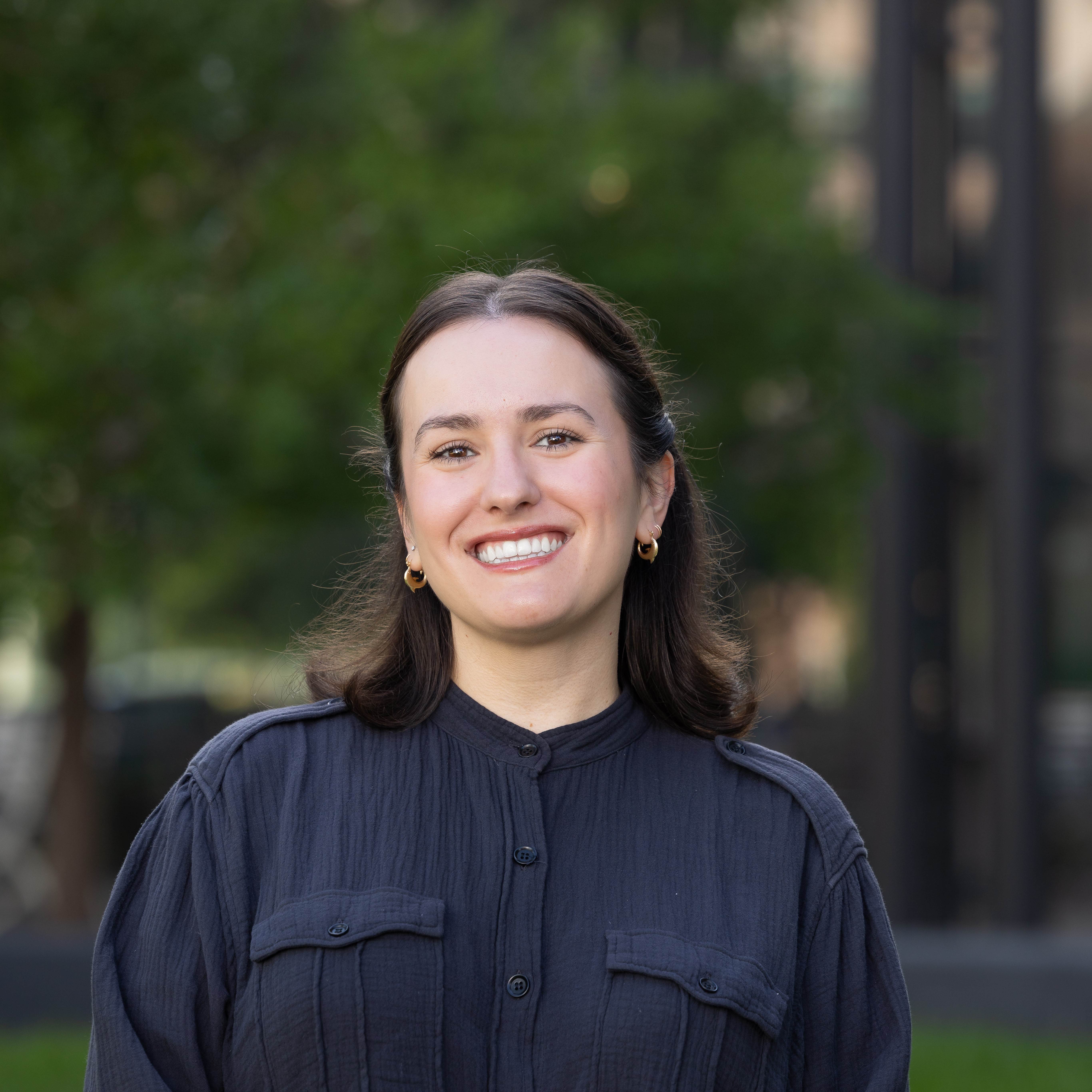 Professional photo of Gillian in front of trees. Gillian is a white woman with straight, short, dark brown hair. Gillian is wearing a black shirt that is buttoned up and gold earrings.