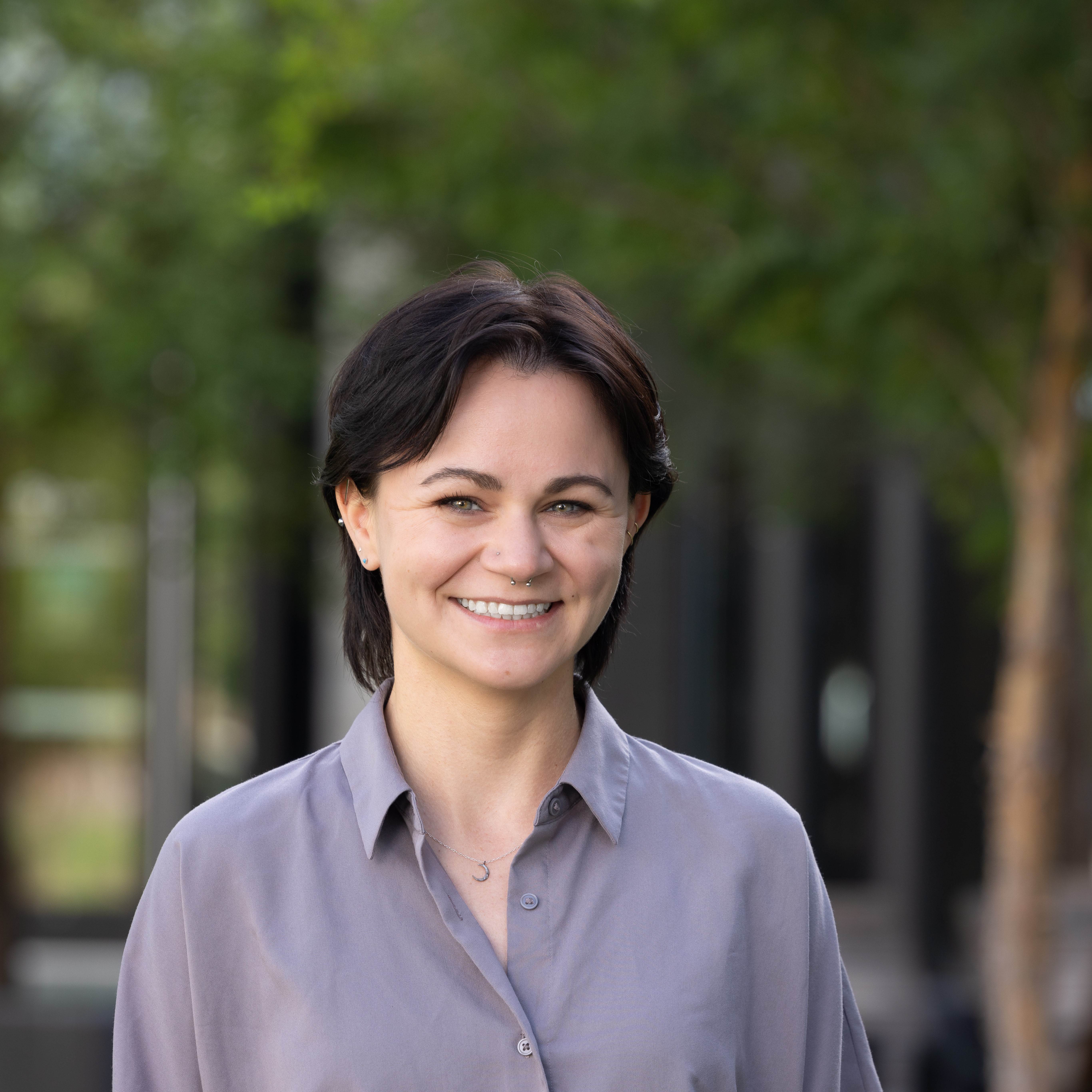 Professional photo of Lindsay in front of trees. Lindsay is a white person with straight, short dark brown hair. Lindsay is wearing a grey shirt that is buttoned up and a crescent moon necklace.