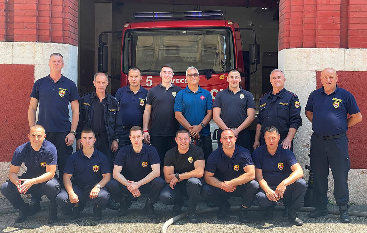 Group of men in navy polo firefighting shirts group together in front of a red truck and fire station.