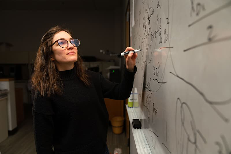 Student in a black sweater and big glasses working on a white board.