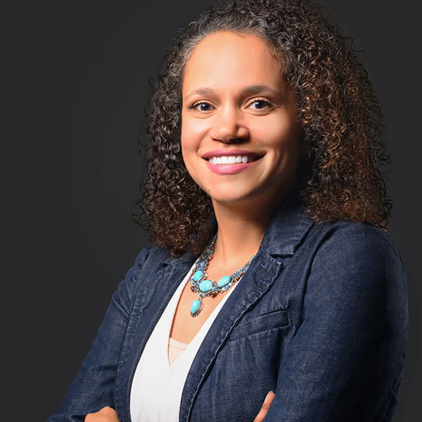 Brandy Reitter in shoulder-length dark, curly hair, wearing a blue suit jacket with her arms crossed, smiling with her teeth.