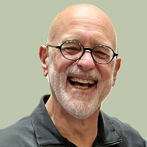 Dan Gilbert in a dark polo, dark-round-rimmed glasses, bald with a short white beard, smiling with his teeth.