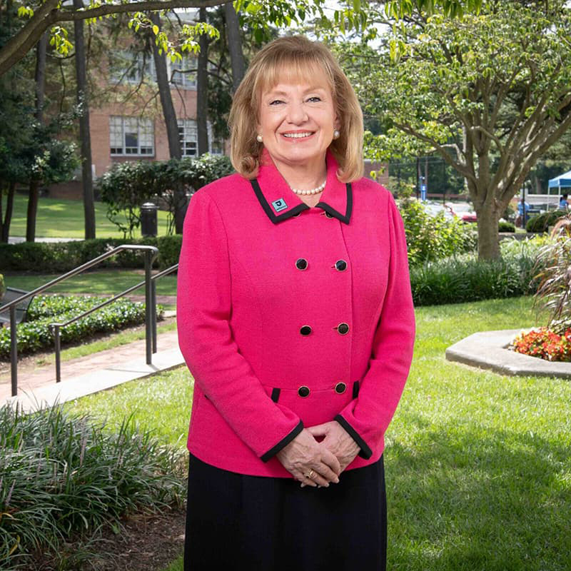 Dr. Susan C. Aldridge standing outside in a pink suit jacket and pearl necklace with shoulder length hair, smiling with her teeth.