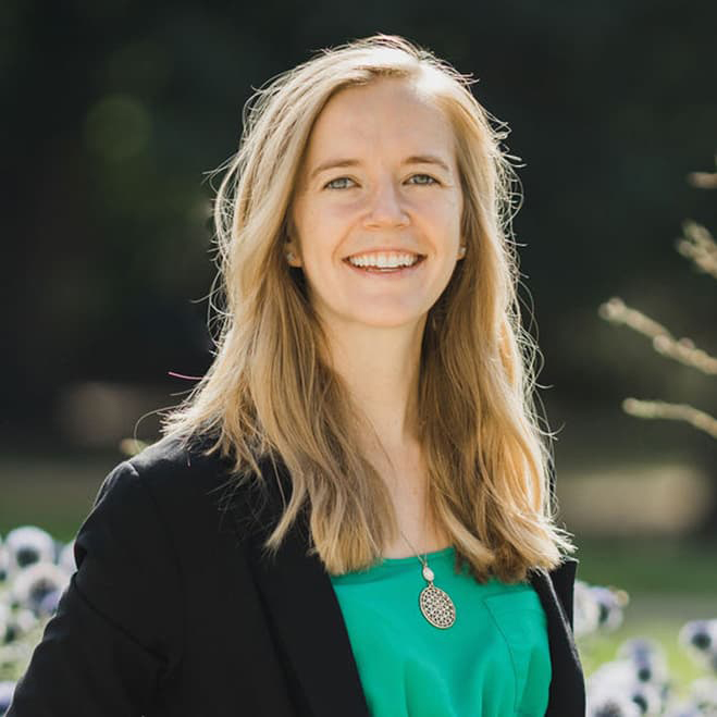 Gabby Hart with a green blouse and black blazer stands outside with long blonde hair moving in the wind and smiling with her teeth.