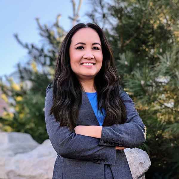 Nga Sandoval with long, dark hair standing outside in a suite with her arms crossed, smiling with her teeth.