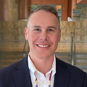 Casey Lozar in a dark suit jacket and white shirt, very short hair, and smiling with his teeth.