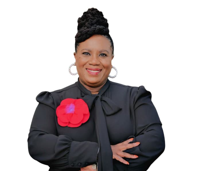 Donna Wilson in a black blouse and a big, brightly colored flower on the chest. She wears pearl hooped earrings and her hair in a detailed bun on top her head. She has a warm smile showing her teeth.