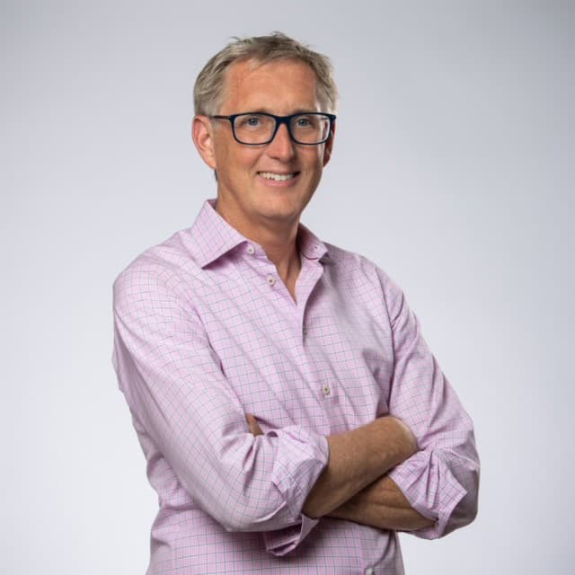 Jamie Van Leeuwen in a pink button-up shirt with arms folded, dark-rimmed glasses, and short, light hair, and smiling with his teeth.