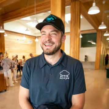 Jeremiah Berndt wearing a dark polo and baseball cap, he has a short beard and smiles with his mouth open.