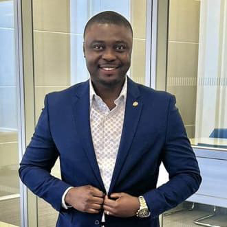 Nagbe Nyenswah in a dark blue suite and short black hair, smiling with his teeth.