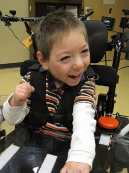 A child with cerebral palsy smiles while seated in his wheelchair. A tray and jelly bean switch are mounted to the chair. 