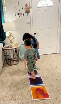 Young brother and sister walk together on a colorful, liquid filled, sensory tile mat on the floor.