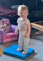 2-year old standing on a blue foam Balance Pad in her family’s living room