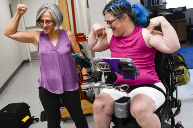 A woman celebrates her wheelchair by flexing her arms alongside a clinician.